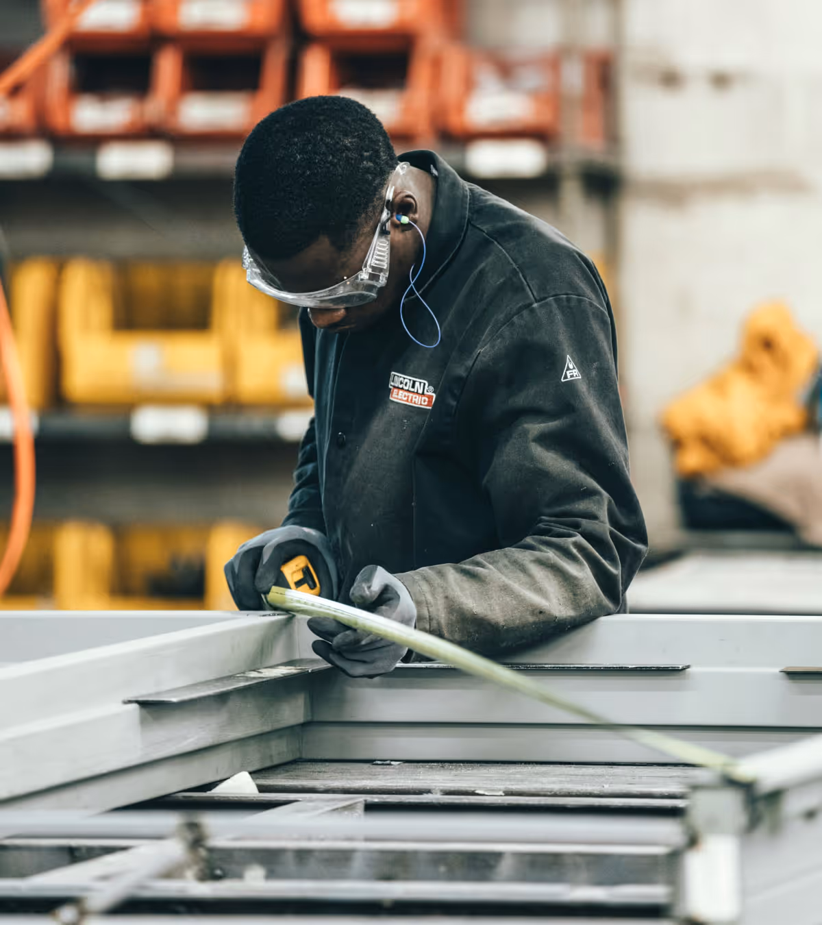 A worker wearing protective goggles and gloves measures a metal frame with a tape measure in an industrial setting.